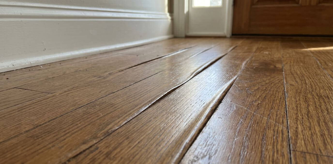 Hardwood floor peaking with raised seams near a wall and doorway