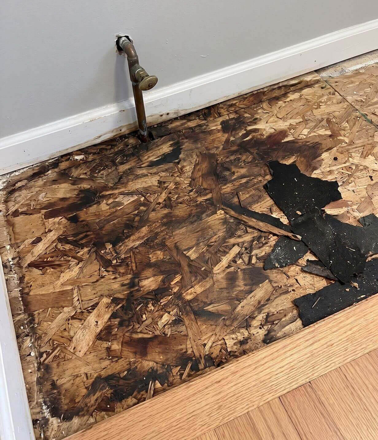 Exposed OSB subfloor with dark moisture staining near a wall pipe